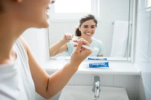 A calm, clean bathroom scene with a person gently brushing their teeth using a soft-bristled toothbrush, a cup of water nearby, and a tube of fluoride toothpaste on the sink. In the background, a mirror reflects a healthy smile. Soft natural light from a window illuminates the scene. No text, no symbols, no medical diagrams, no visible wounds or inflammation in the mouth.