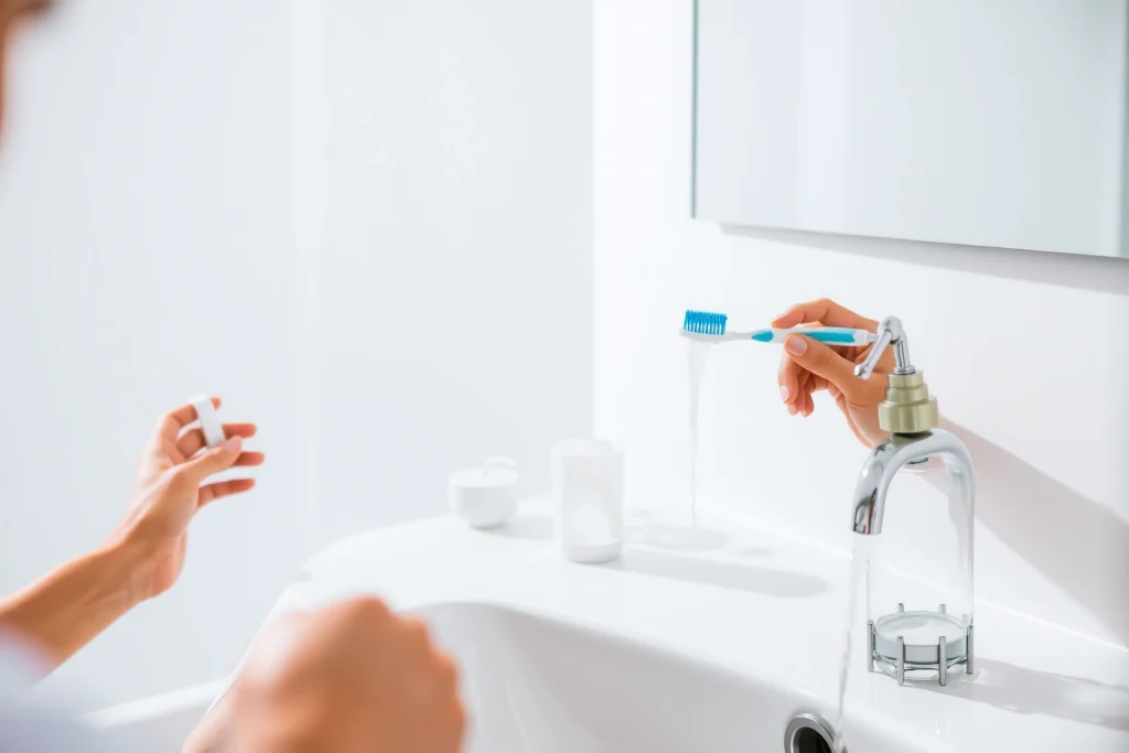 A clean, modern bathroom with a person brushing their teeth using a toothbrush and toothpaste, a dental floss container, and a cup of water flowing from a faucet, soft natural lighting, realistic details, no text or symbols visible