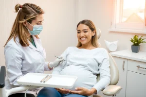 A pregnant woman smiling gently while sitting in a modern dental chair, receiving a routine check-up from a kind female dentist wearing a mask and gloves, with soft natural light coming through a window, dental tools neatly arranged on a tray, a small potted plant on the counter, warm and calming atmosphere, no text or symbols visible