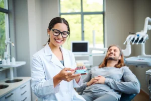 A calm, modern dental clinic interior with a smiling dentist in a white coat gently holding a dental implant model, a patient relaxing in the dental chair with a reassuring expression, soft natural light from large windows, subtle medical equipment in the background, warm and inviting atmosphere, no text or symbols visible