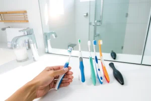 A clean, modern bathroom setting with a person's hand holding a toothbrush, showing different types of toothbrushes (manual, electric, soft bristles, hard bristles) arranged neatly on a white countertop, with a mirror in the background reflecting soft natural light, emphasizing oral hygiene and proper selection