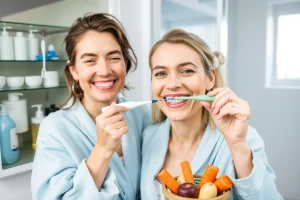 A cheerful adult woman in a bright, modern bathroom, wearing a light blue robe, brushing her teeth with a white toothbrush and a mint green toothpaste tube. She is smiling, showing healthy, white teeth. In the background, a glass cabinet displays a variety of dental hygiene products: floss, mouthwash, a dental mirror, and a small jar of sugar-free gum. A healthy fruit bowl with apples and carrots sits on a nearby counter, symbolizing good nutrition. The scene conveys a clean, hygienic environmen