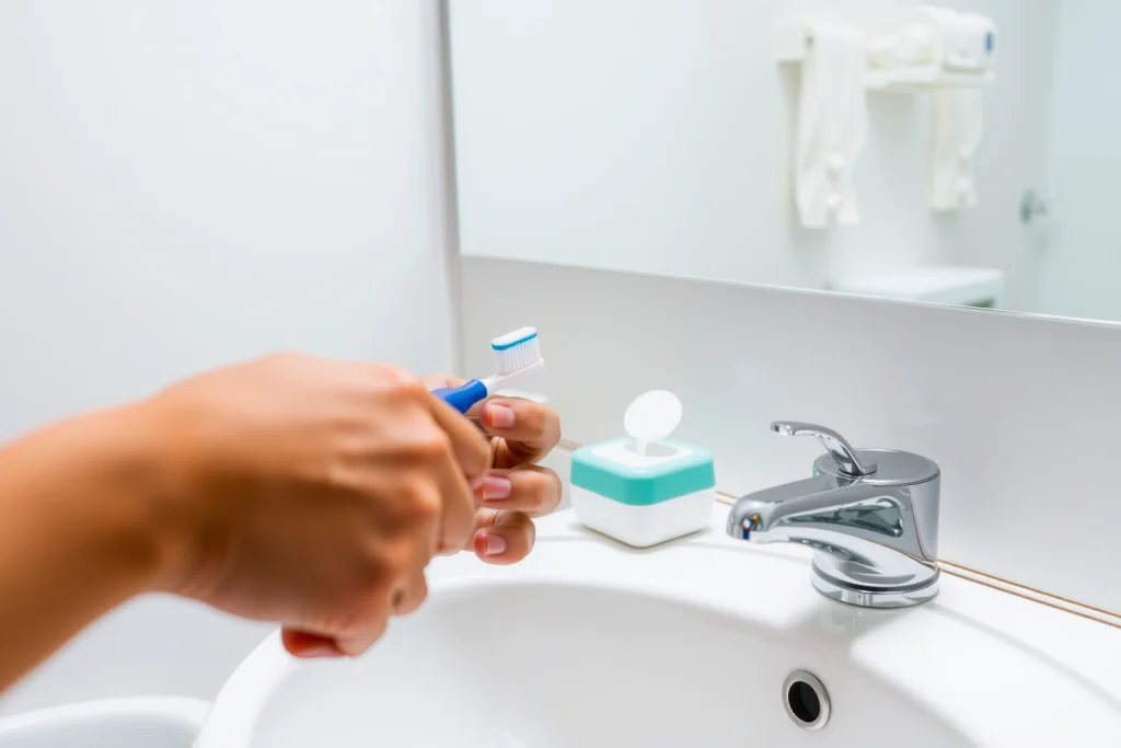 A clean, modern bathroom with a person brushing their teeth using a toothbrush and toothpaste, a dental floss container, and a cup of water on the sink, soft natural lighting, realistic details, no text or symbols