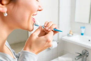 A person brushing their teeth with toothpaste on a toothbrush, showing proper technique with gentle circular motions, in a clean modern bathroom setting with a mirror and sink visible in the background, soft natural lighting, focus on the hands and toothbrush, no text or labels